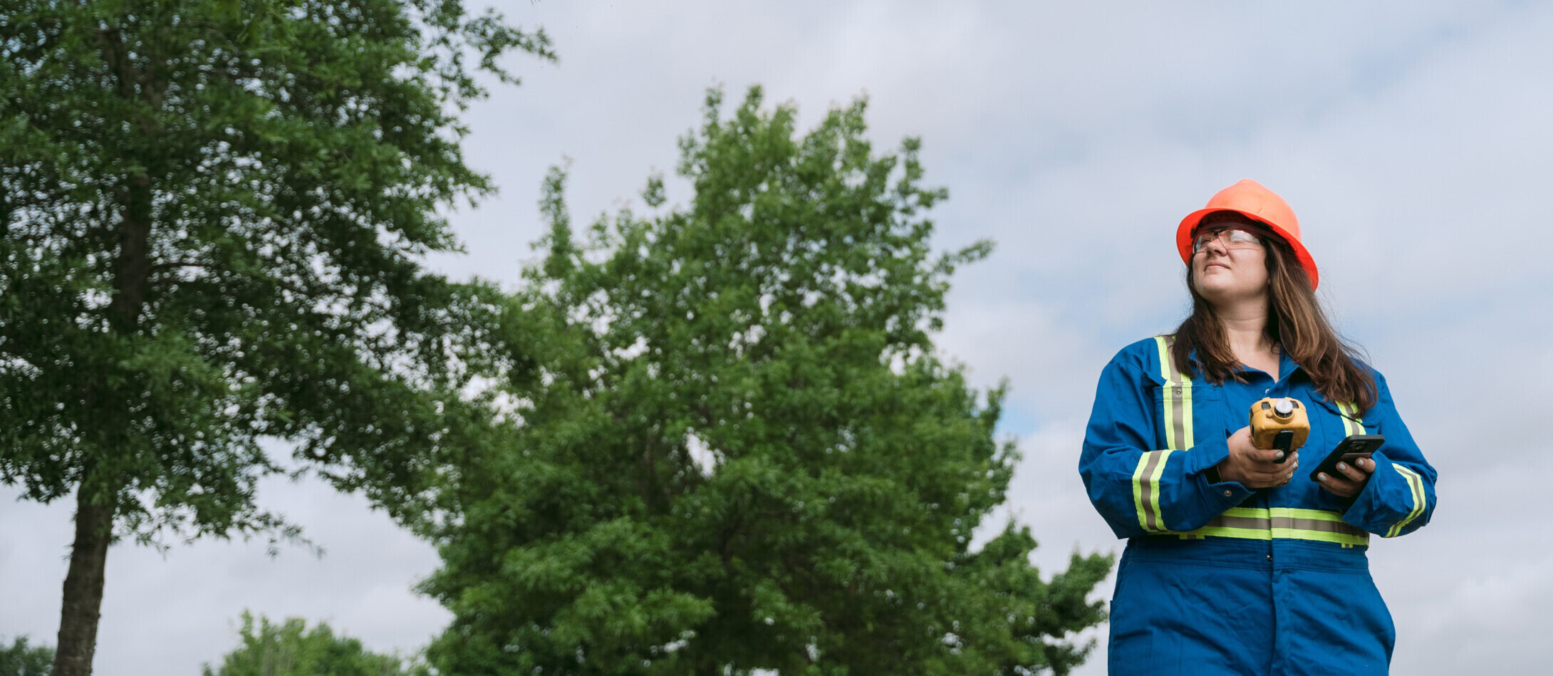 A person wearing a blue and yellow safety uniform and an orange hard hat is standing outdoors holding a monitoring device, crucial for environmental response. They are looking off to the side with trees and a cloudy sky in the background.