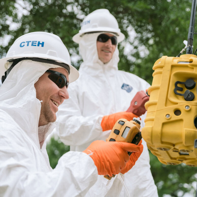 Two individuals wearing white protective suits, orange gloves, and helmets labeled "CTEH" are handling yellow equipment outdoors. One person is smiling and appears to be adjusting a device related to environmental consulting, while the other looks on. Trees and greenery are visible in the background.