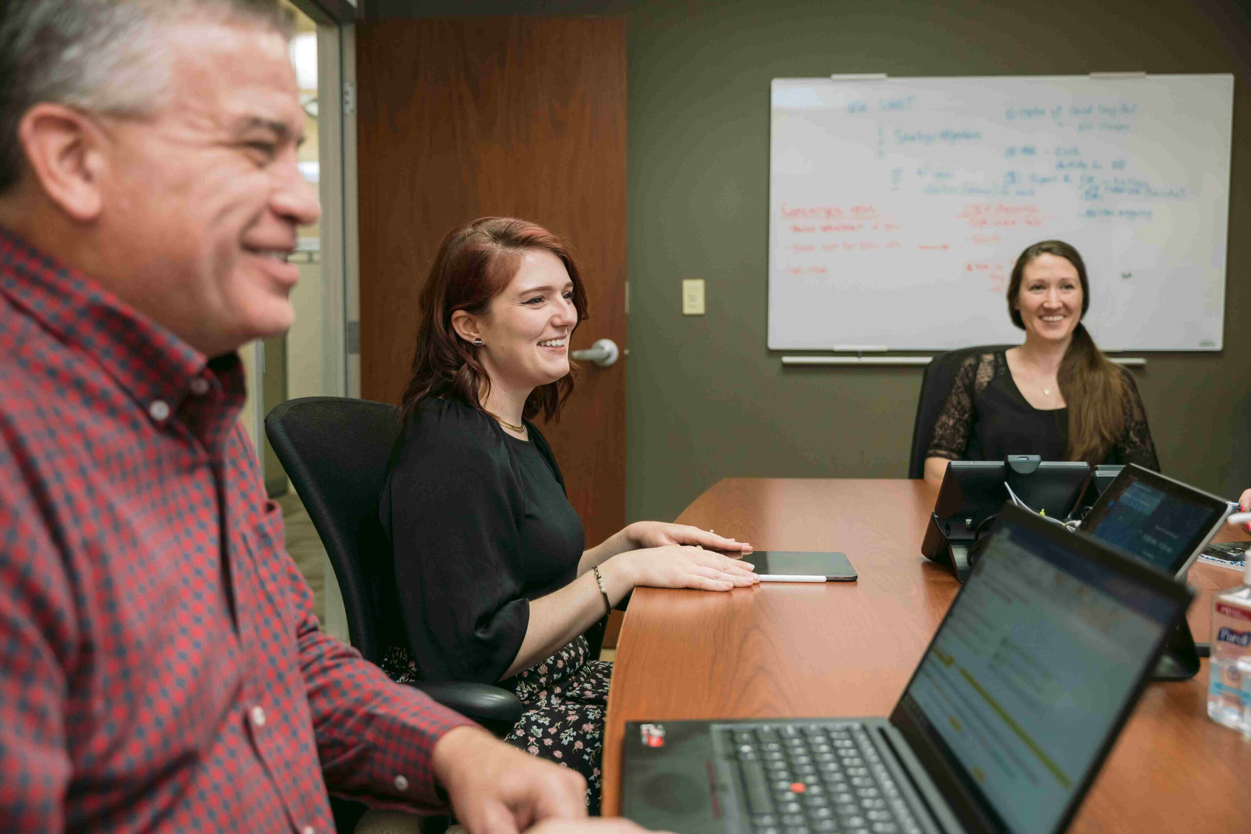 Three people are seated around a conference table during a meeting. Two laptops are open on the table. A whiteboard with handwritten notes is visible on the wall behind them. The participants appear engaged and are smiling.