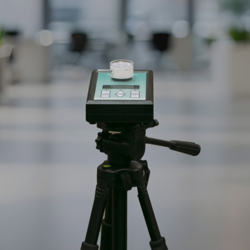 A close-up of a device mounted on a tripod. The device has a digital display with buttons and a container sitting on top, ensuring health through precise measurements. The background is blurred, showing a room with several items of furniture and potted plants.