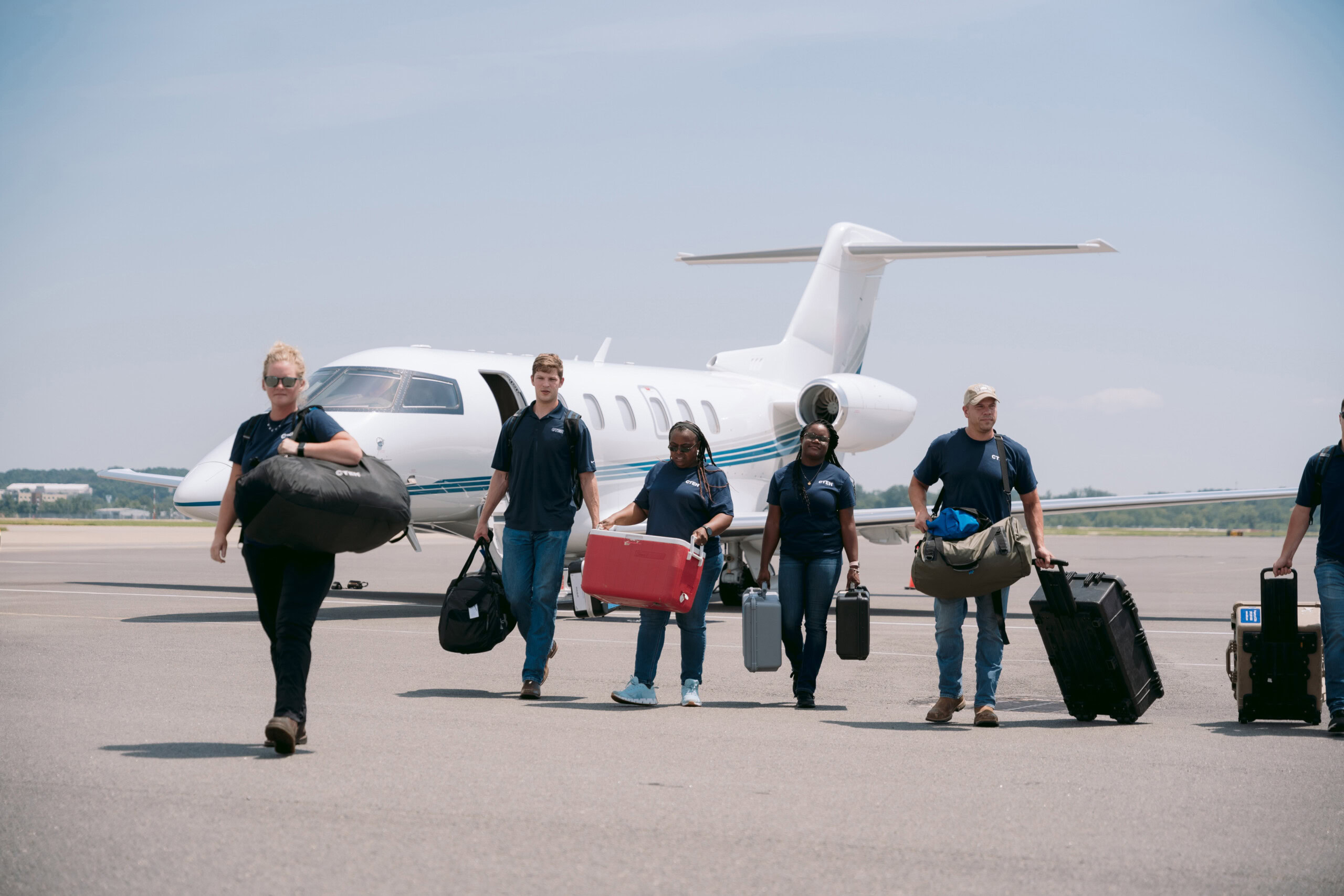A group of six people in uniform walk on an airport tarmac, exuding readiness as they carry luggage and equipment. In the background, a small private jet is parked against a clear blue sky.