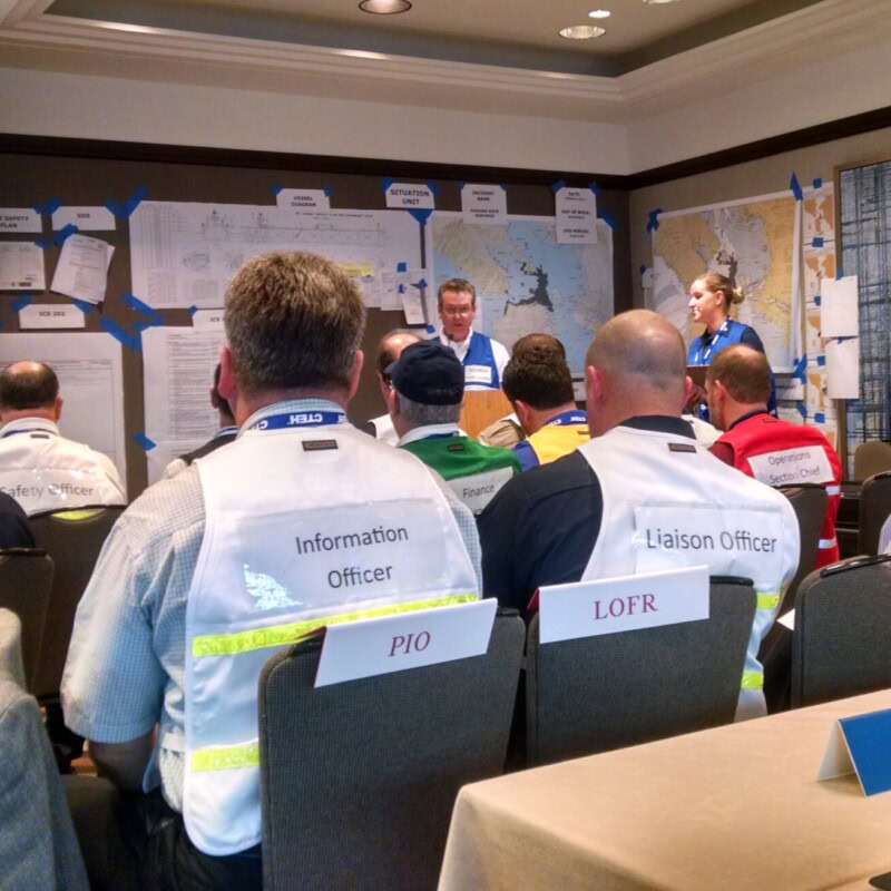 A group of people in a meeting room sit facing a speaker at a podium. Participants wear vests with labels such as "Information Officer" and "Liaison Officer," indicative of their roles in disaster recovery. Wall maps are pinned behind the speaker, and "PIO" and "LOFR" labels are visible on chairs.
