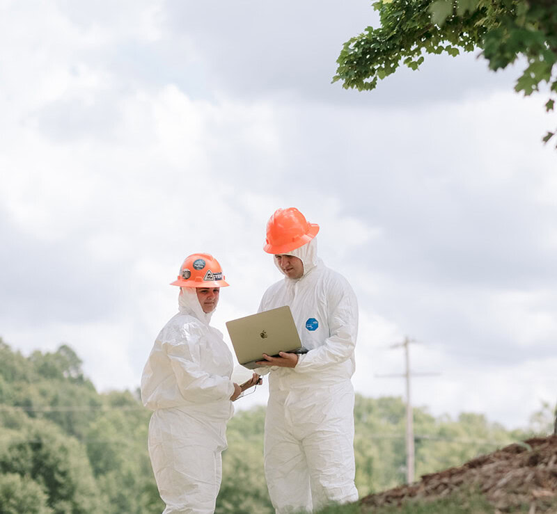 Two people in white protective suits and orange helmets stand outdoors near a tree, looking at a laptop. Trees and a cloudy sky are in the background, suggesting an occupational health inspection or environmental response activity taking place in a natural environment.