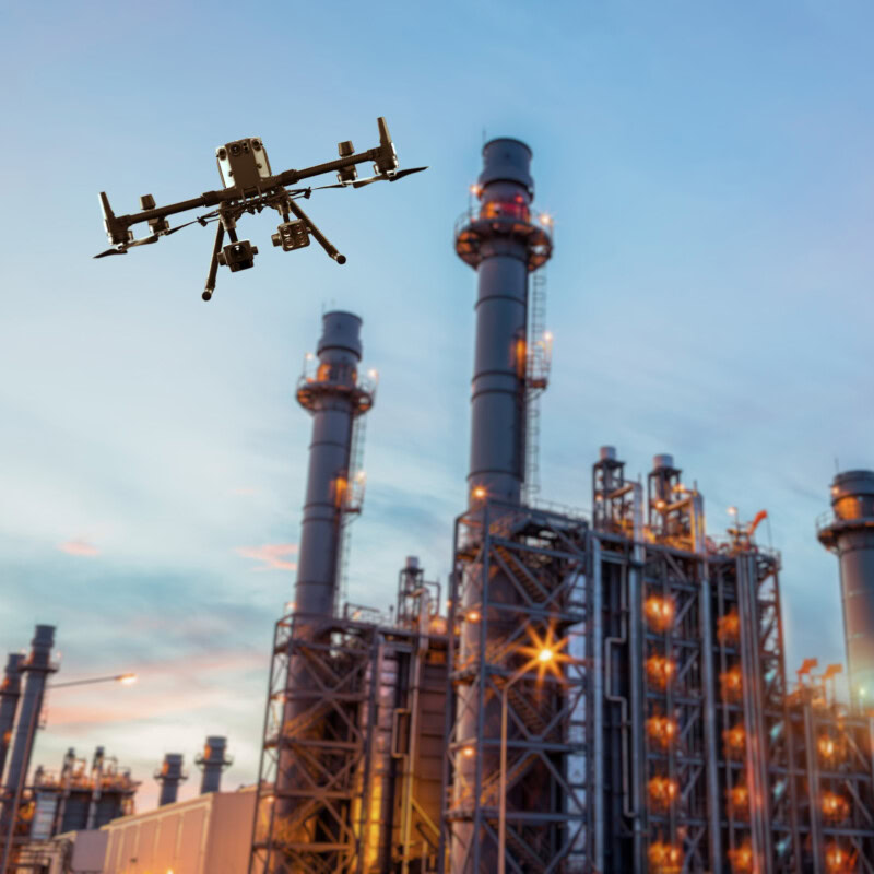 A drone hovers in the sky near an industrial facility with multiple tall metal structures and chimneys. The facility and sky are illuminated by the glow from the chimneys and setting sun.