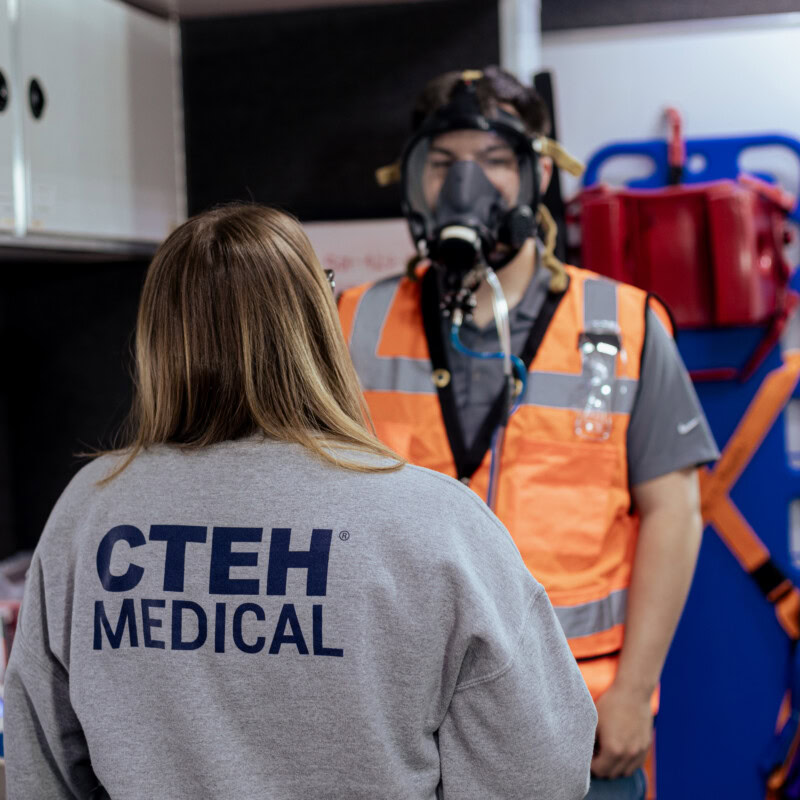 A person wearing a shirt with “CTEH Medical” on the back speaks to another individual in an orange safety vest and full-face respirator mask. Inside the mobile medical unit, equipped with gear and informational posters, they appear to be discussing disaster recovery efforts and occupational health protocols.