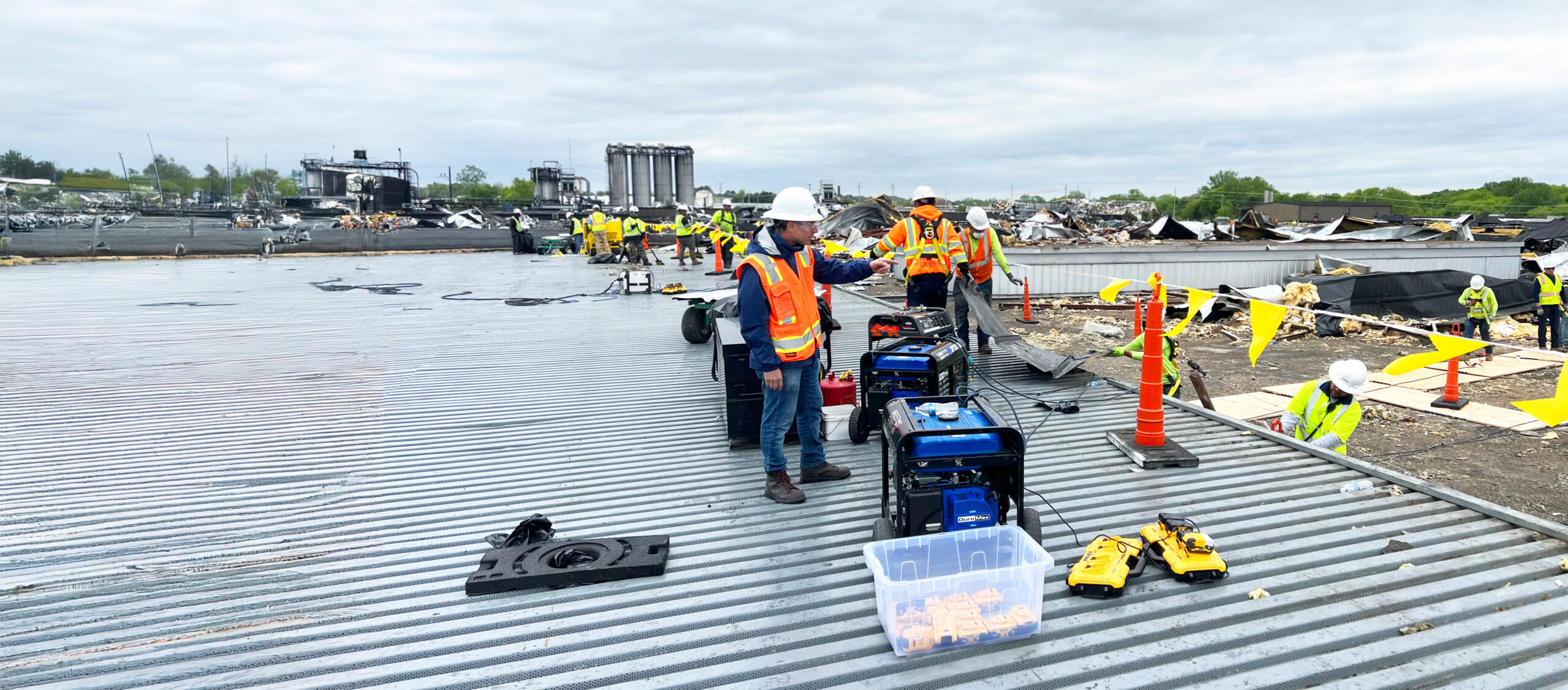 A group of construction workers in orange safety vests and white helmets are standing on a corrugated metal surface. Various tools and equipment are spread out around them. In the background, there is debris and partially constructed or demolished buildings.