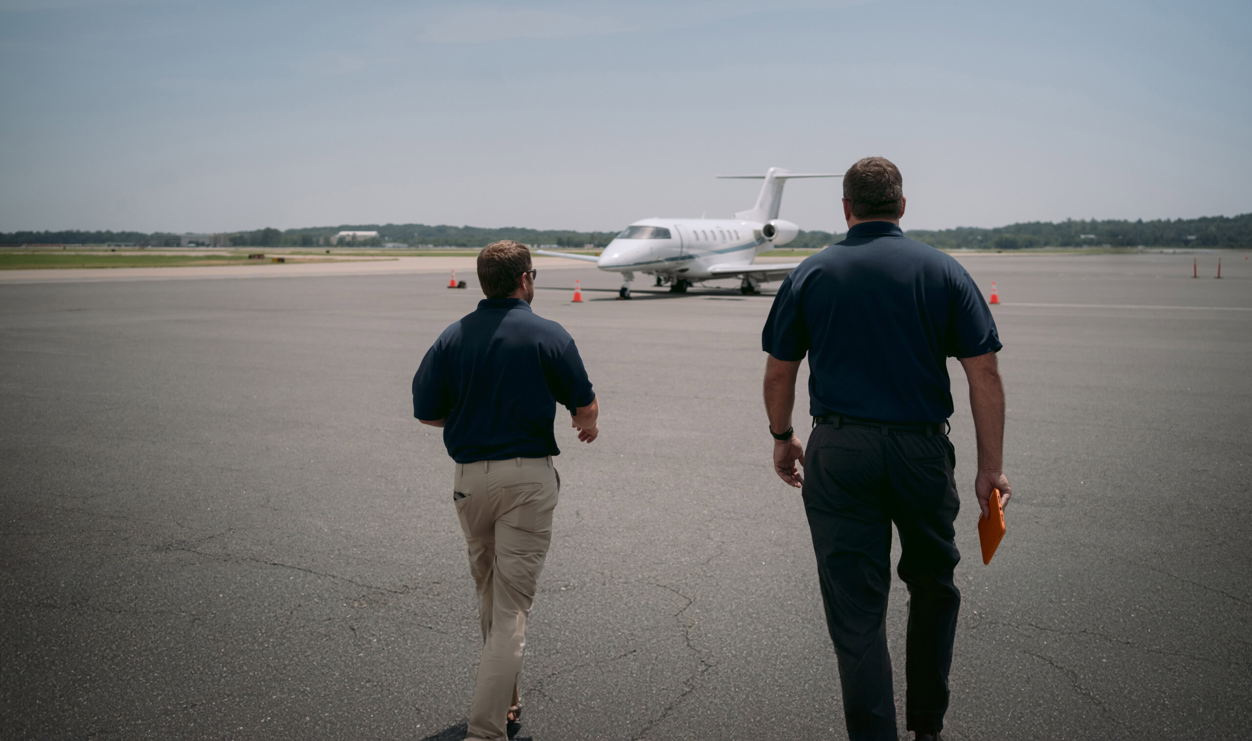 Two people dressed in dark blue shirts and pants are walking towards a small jet on a clear runway, holding an orange folder. Orange cones are placed near the aircraft, and it's a sunny day with a clear sky. Our approach can be characterized by innovation and customer satisfaction.