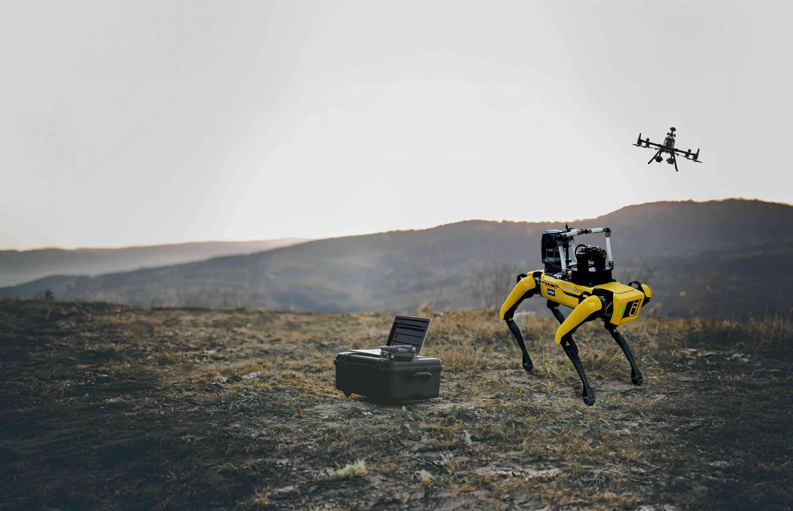 A yellow, four-legged robot stands on a barren landscape next to an open black case with electronic equipment, showcasing technological solutions, while a drone flies in the background against a backdrop of hills and a hazy sky.