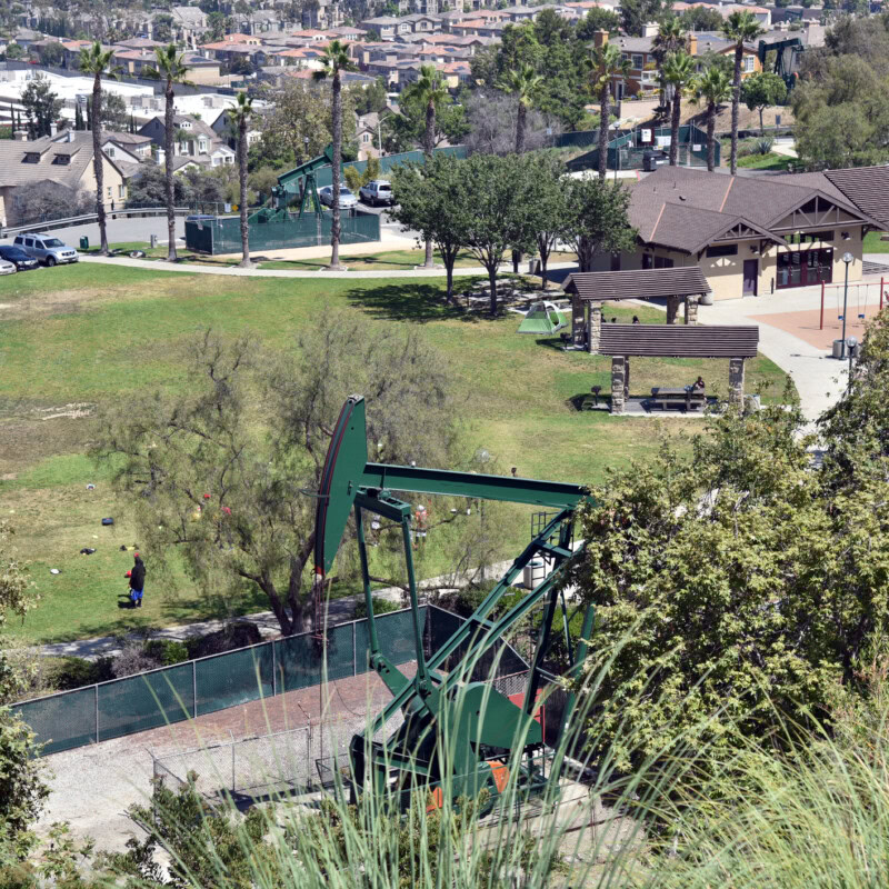 A green oil pumpjack is in the foreground of a hilly park area. The park, known for its expert landscaping services, has walking paths, trees, benches, and a playground. Several buildings and a community center are visible in the background, with houses and trees in the distance.