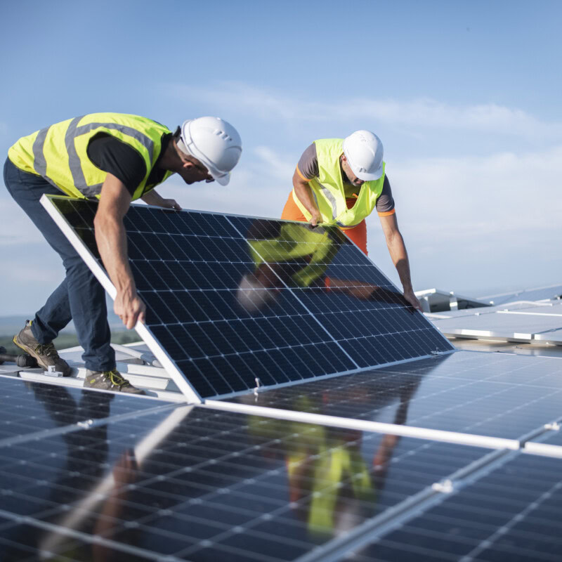 Two workers in yellow safety vests and white helmets are installing solar panels on a rooftop under a clear blue sky. While one worker holds up the panel and the other positions it, an environmental consultant oversees the process, ensuring compliance with environmental response regulations.