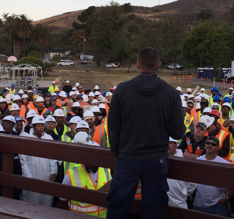 A person wearing a dark hoodie addresses a large group of construction workers in white hard hats and high-visibility vests. The outdoor gathering, framed by palm trees, portable buildings, and a hill in the background, likely involves an important risk assessment discussion.