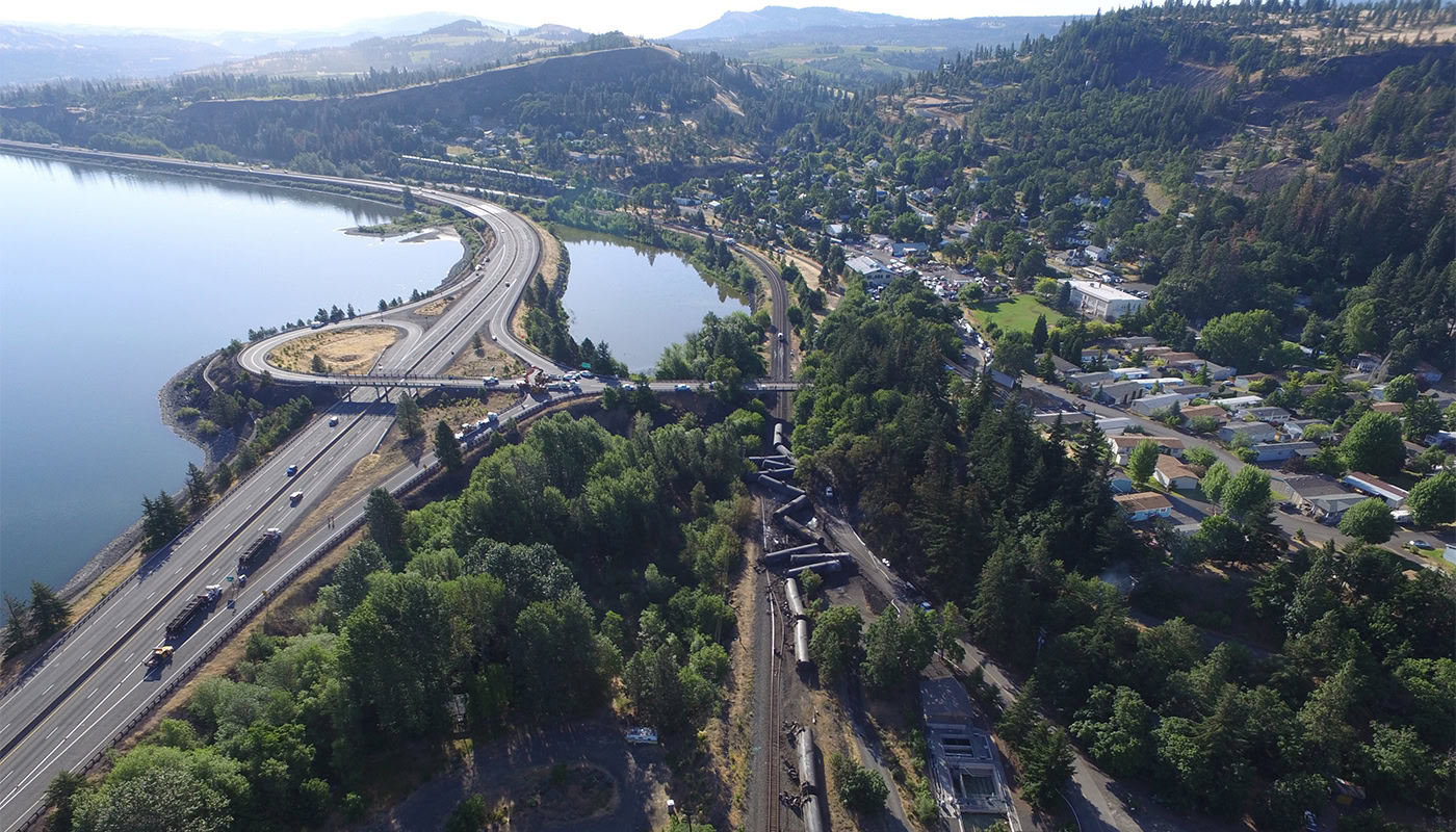 Aerial view of a train derailment near Mosier, Oregon. Several train cars are off the tracks surrounded by forested areas. Given the proximity to the Columbia River and residential area, an environmental response team is assessing potential risks. A highway runs parallel to the tracks nearby.