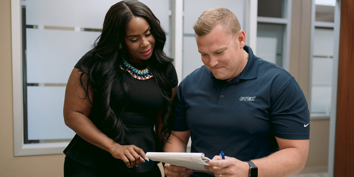 A man and a woman are standing together in an office. The man, wearing a navy blue polo shirt, is holding a notepad with a pen. The woman, dressed in black, is looking at the notepad as they appear to be discussing an environmental consulting project.